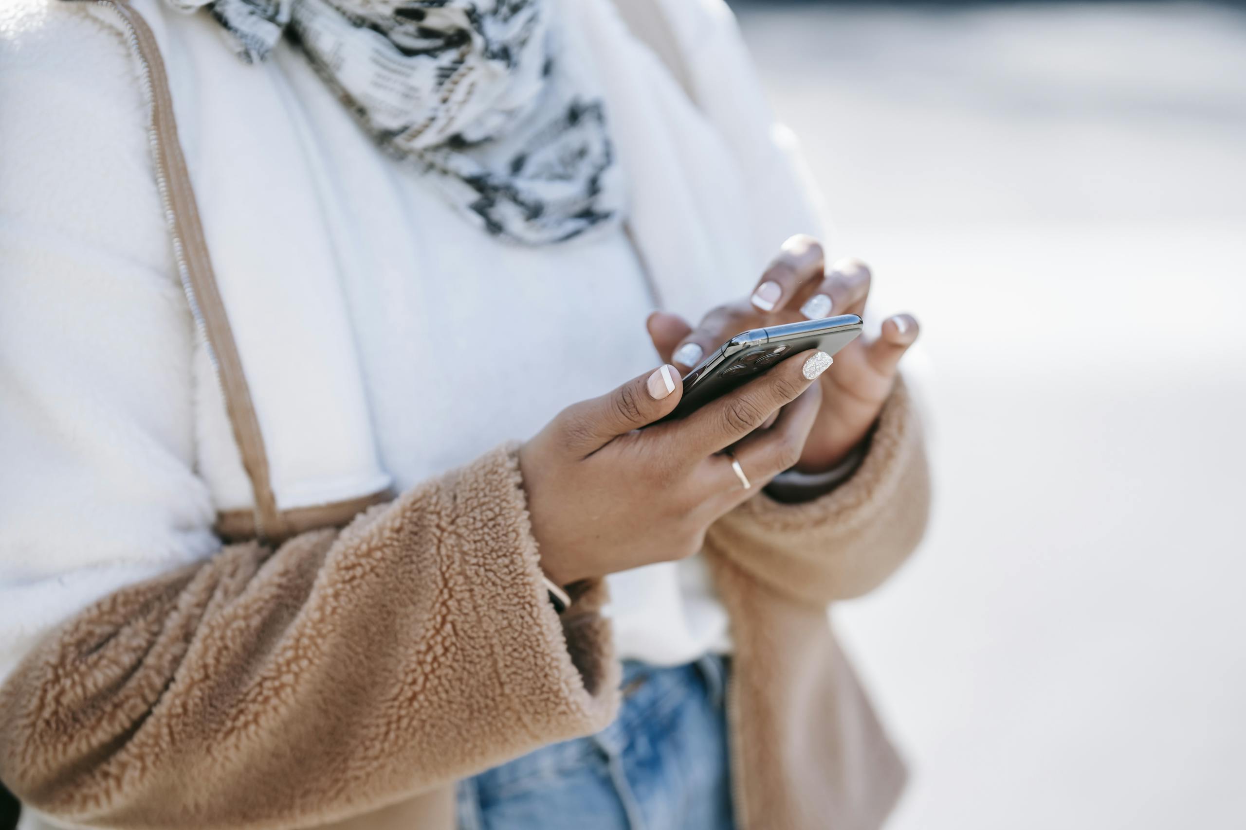 Crop anonymous woman wearing fluffy warm jacket and scarf using contemporary smartphone while standing outdoors